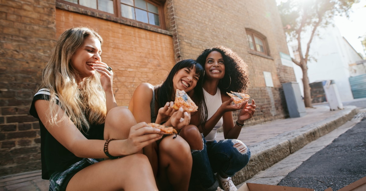beautiful girls sitting on pavement eating pizza together