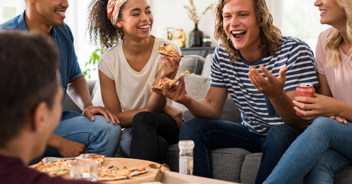 friends sitting on couch eating pizza while talking to each other at hose party