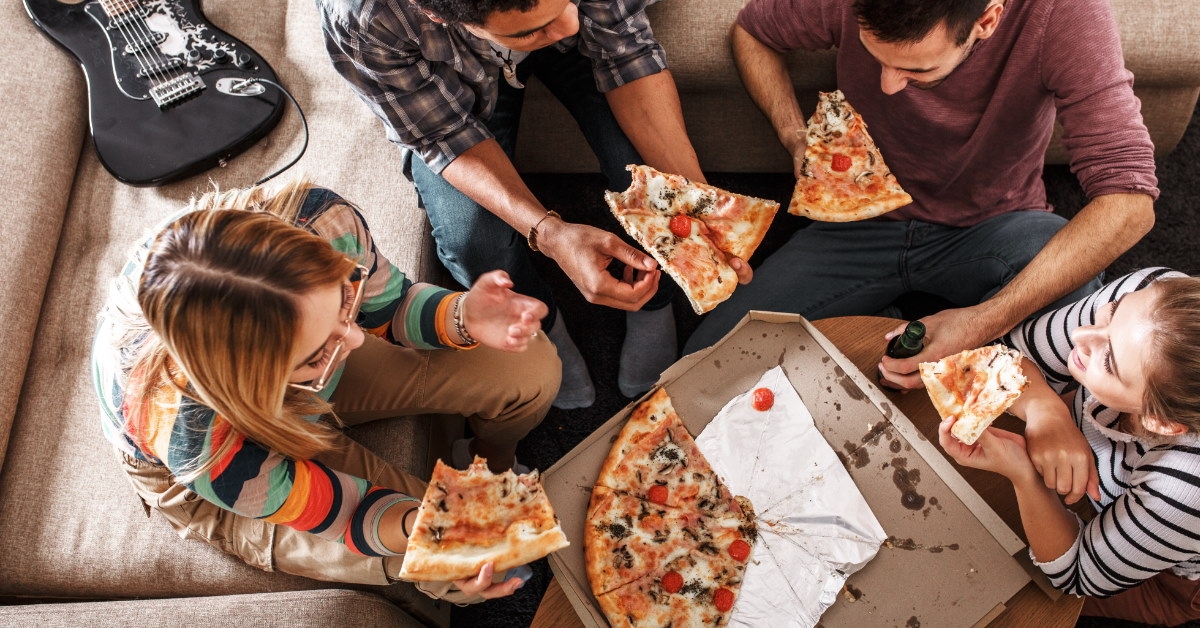 friends eating pizza on couches in home lounge with pizza box on table.