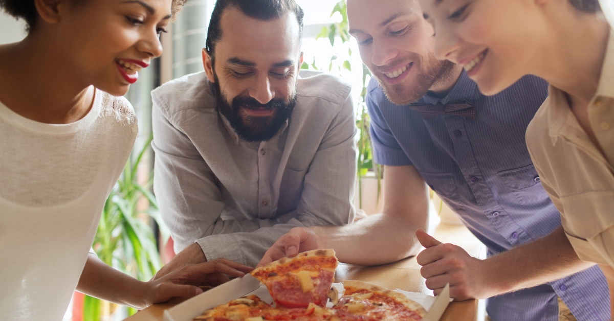 group of happy friends sitting together at table eating pizza from box at office