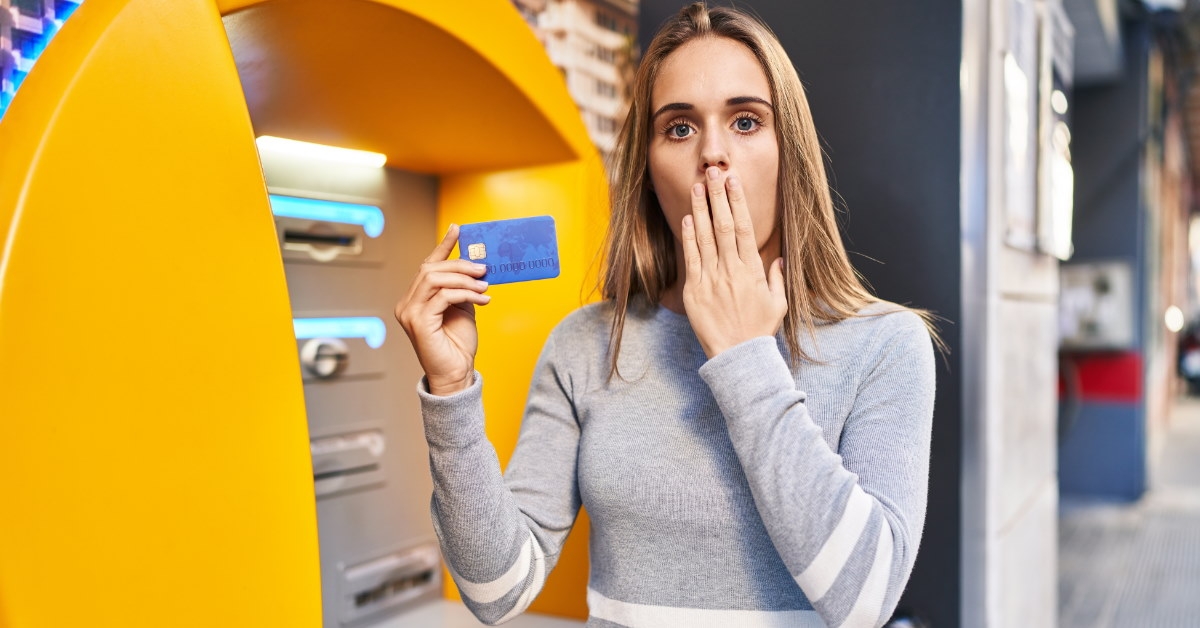 woman standing at atm covering mouth in shock using hand while showing card