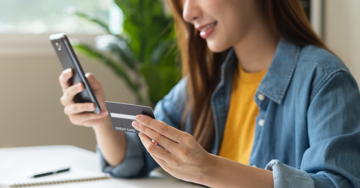 happy woman sitting at table holding card to shop using smartphone