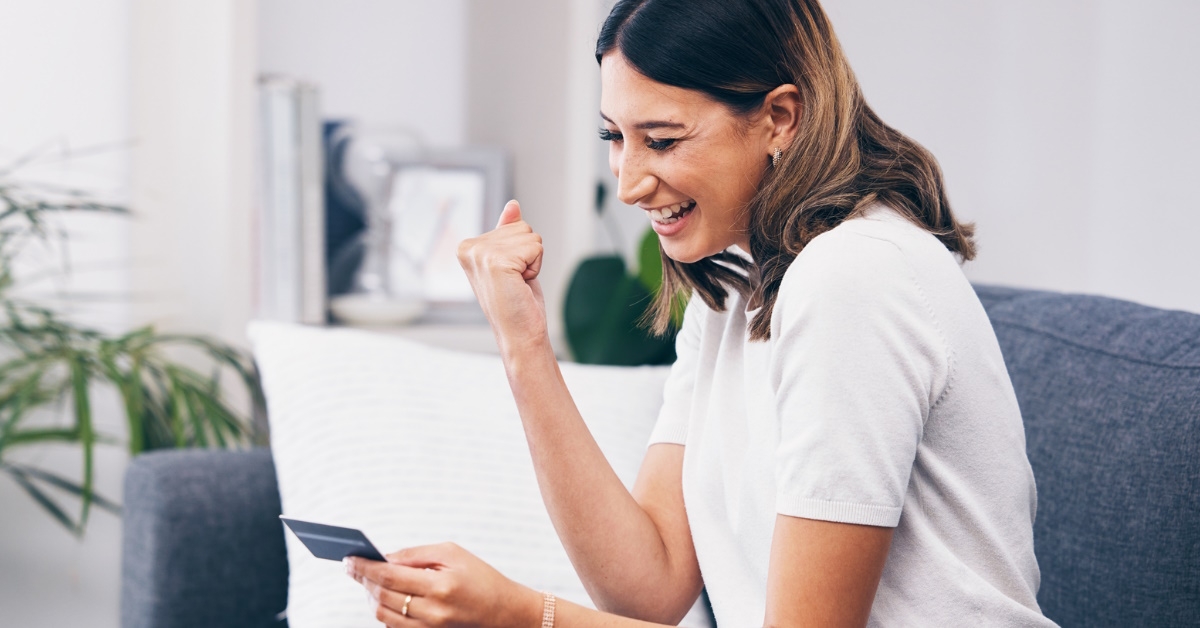 woman sitting on couch at home celebrating while using laptop and holding card in hand