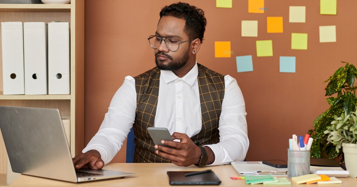 african american man sitting at table using laptop for work while holding smartphone