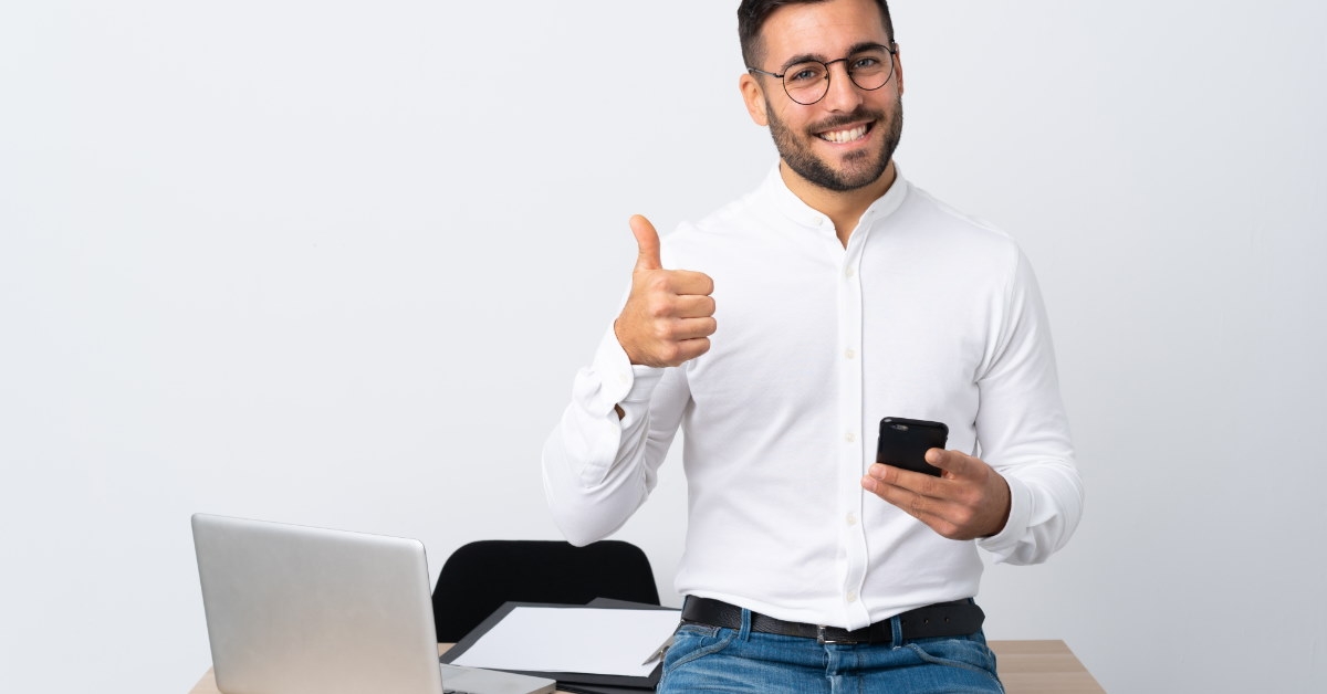 man standing in front of table doing thumbs up sign while using smartphone at work