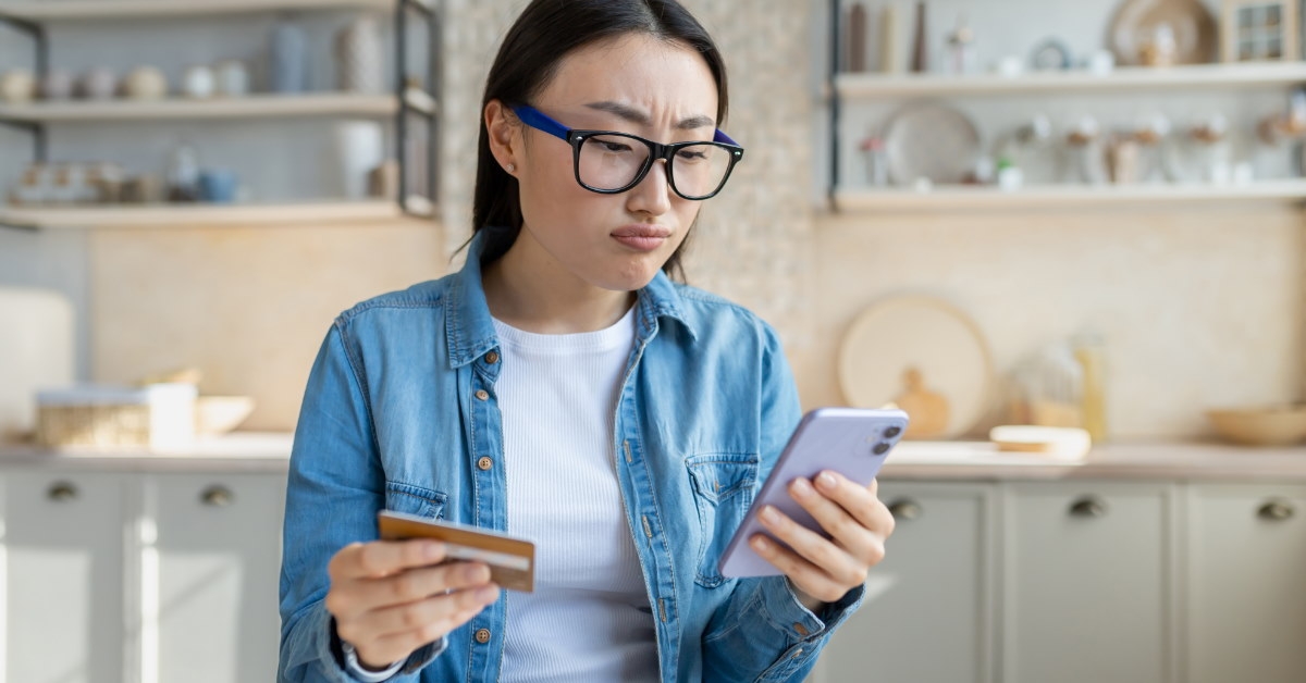 upset asian woman sitting at table in kitchen looking at smartphone screen while holding card