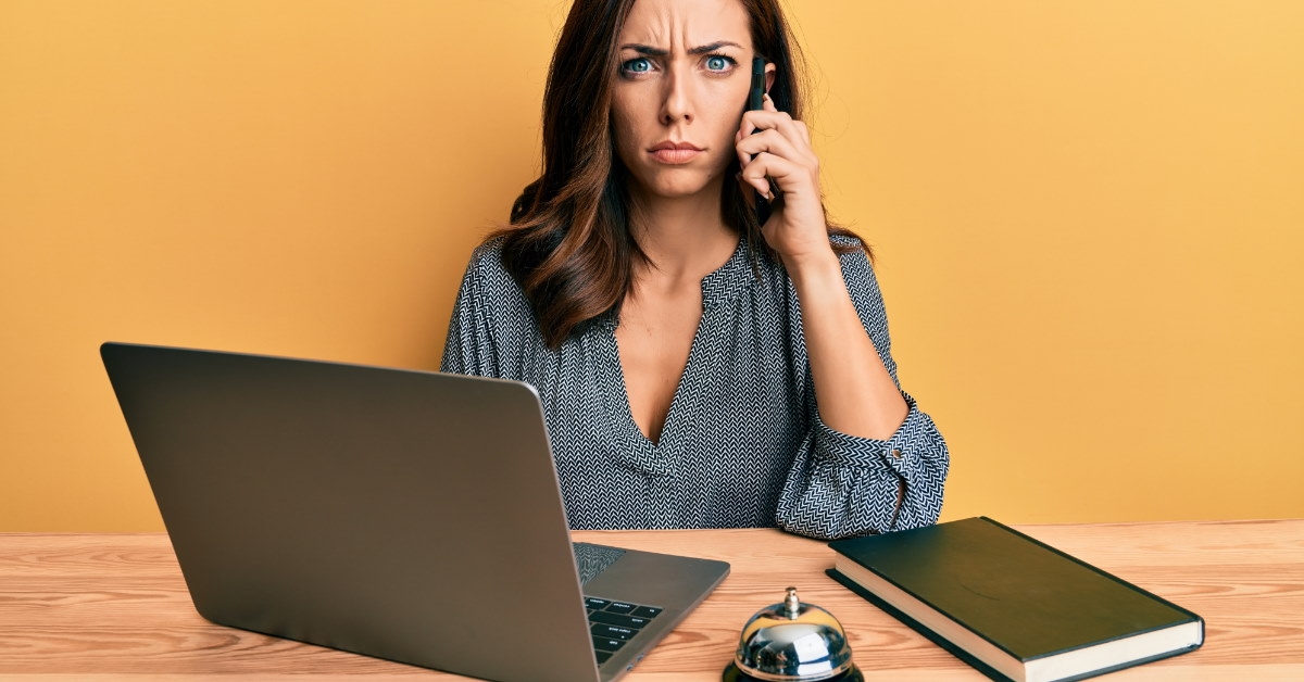woman sitting at table with book and laptop frowning while talking on smartphone