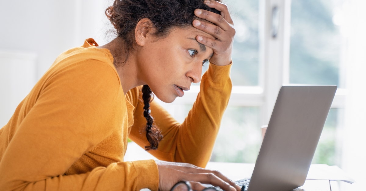 woman sitting at table holding head in stress while using laptop