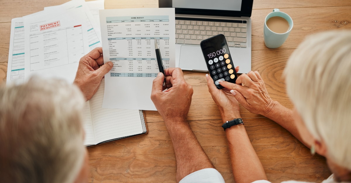 senior couple reviewing bills at table while using calculator on smartphone with laptop 