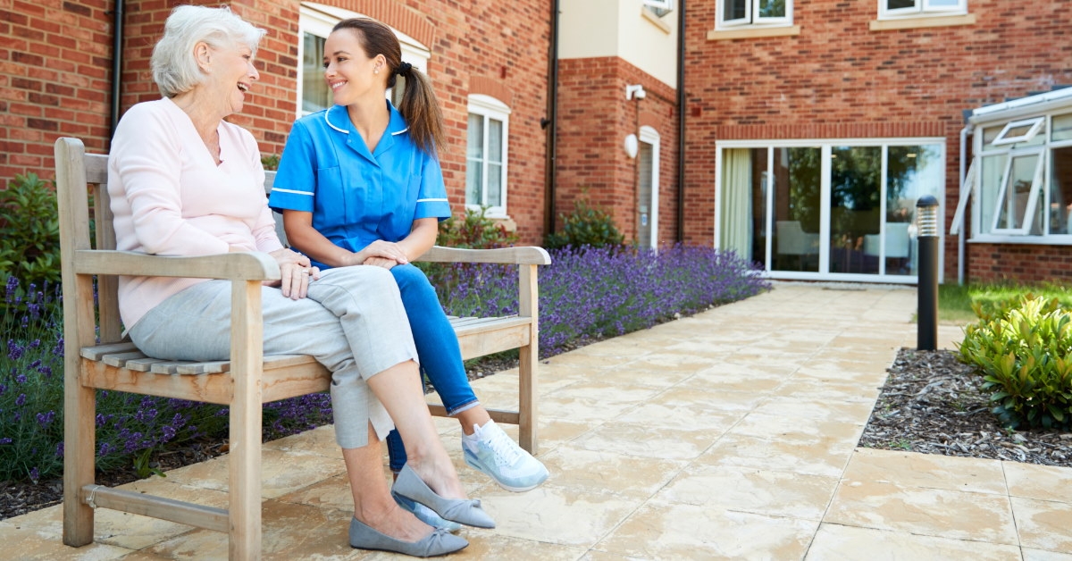 Senior woman sitting on bench with female nurse at nursing home during day time