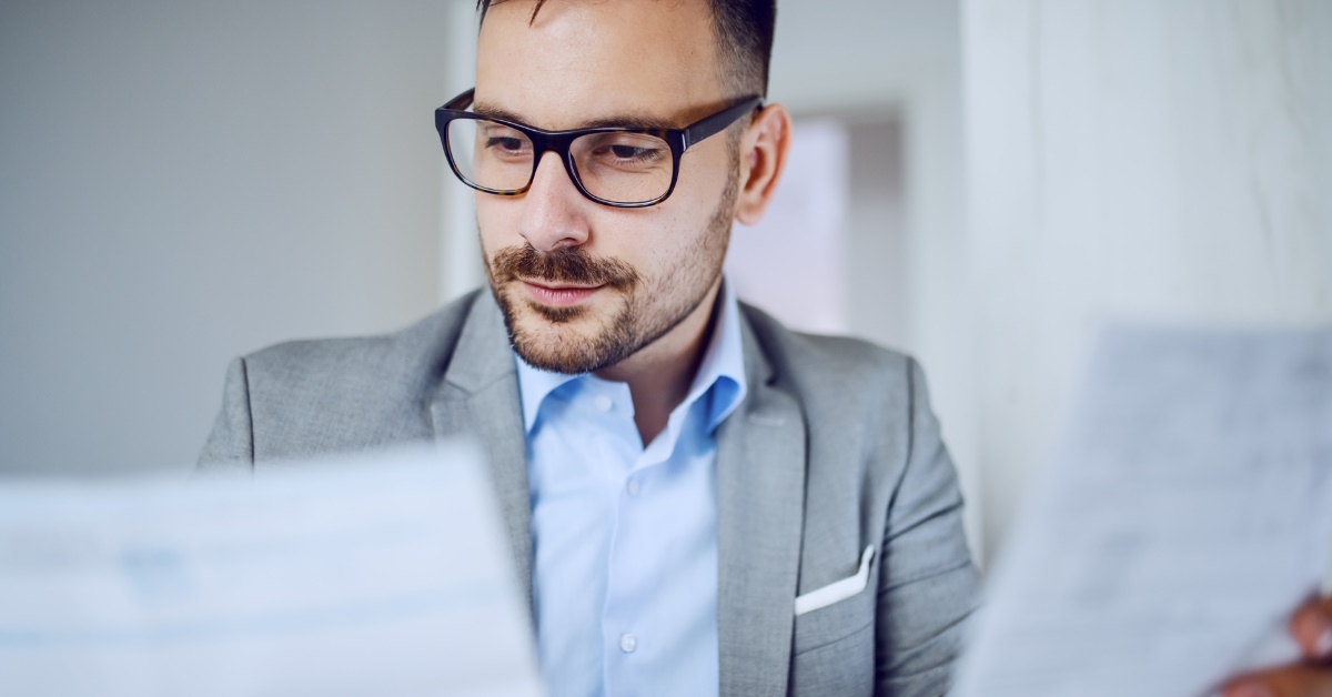 businessman wearing suit and glasses sitting at office reviewing documents in hand