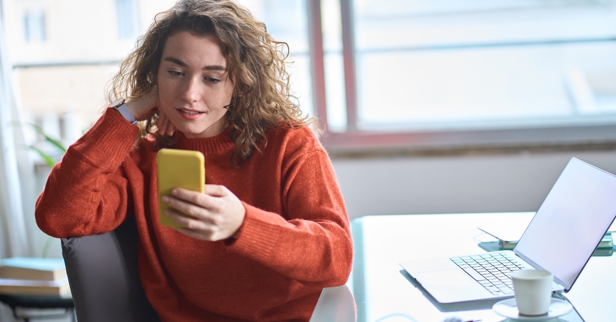 young woman sitting at office wearing jumper scrolling through smartphone in hand with laptop at table