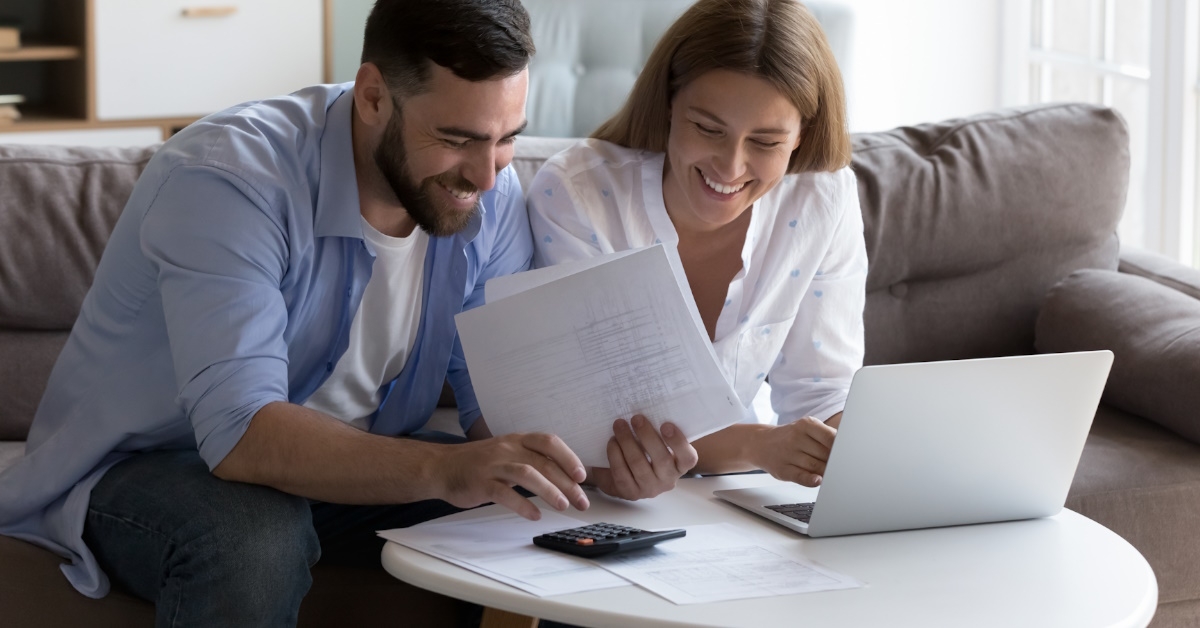 happy couple sitting on couch at home using laptop while reviewing bills 