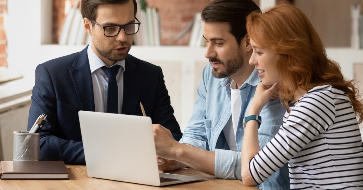male real estate agent showing plans on laptop to young couple at office