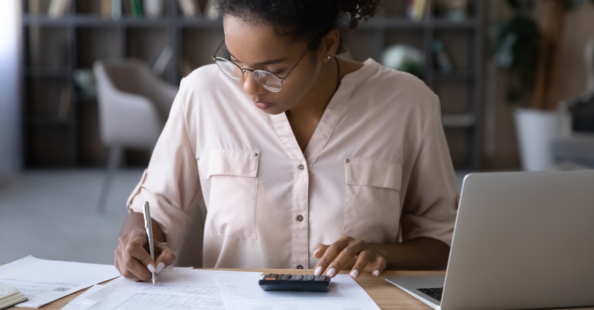 african american woman calculating budget at table while writing expenses besides laptop