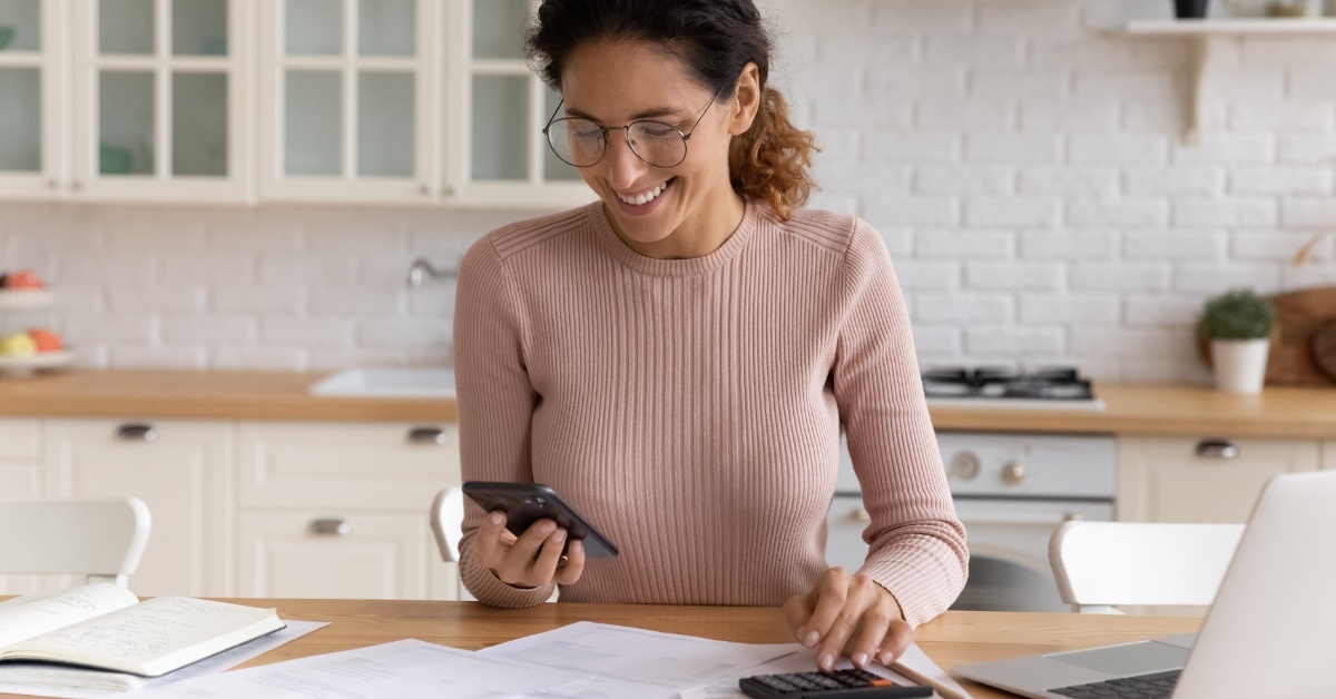 happy woman sorting out budget in kitchen while holding smartphone