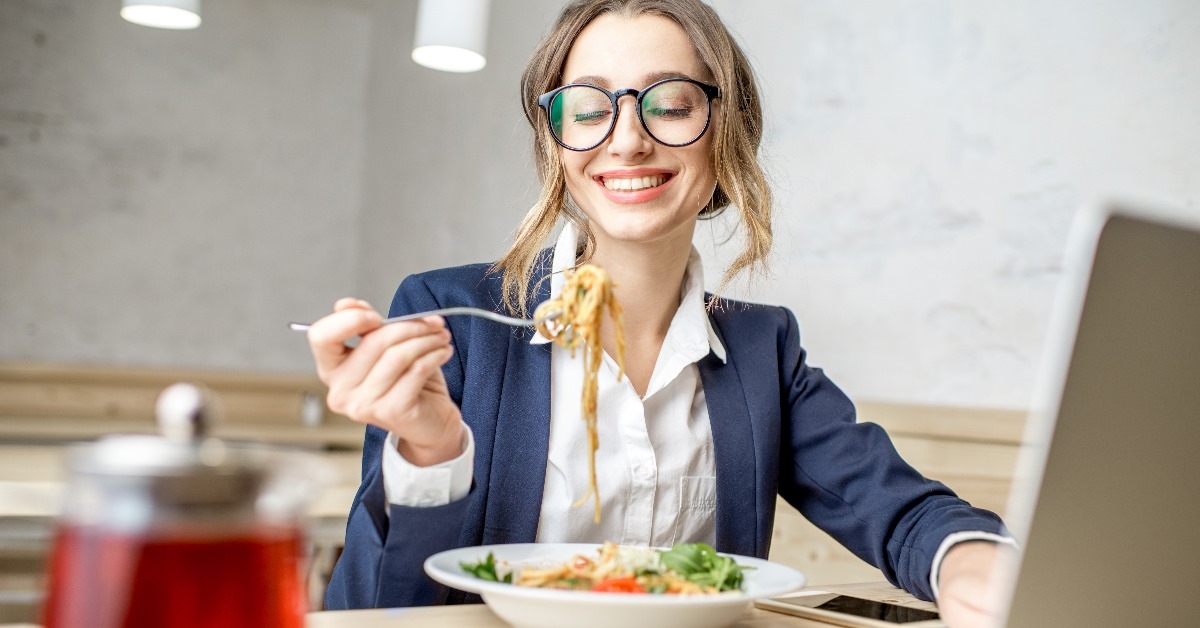 businesswoman sitting at table with laptop eating pasta in lunch at office