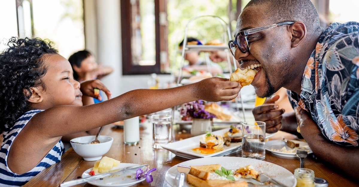young daughter feeding her father bread at a restaurant.
