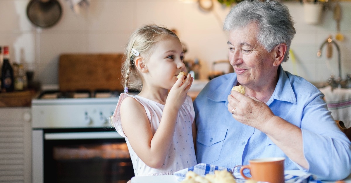 senior woman sitting on chair at kitchen enjoying cookies with small girl