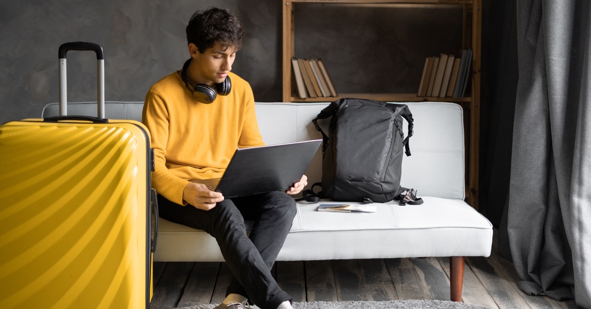 male tourist sitting on couch in hotel room using laptop with yellow suitcase besides him