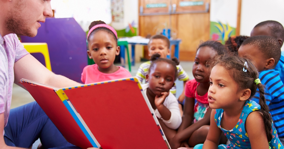 male volunteer sitting on ground reading out from book to class of african american kids