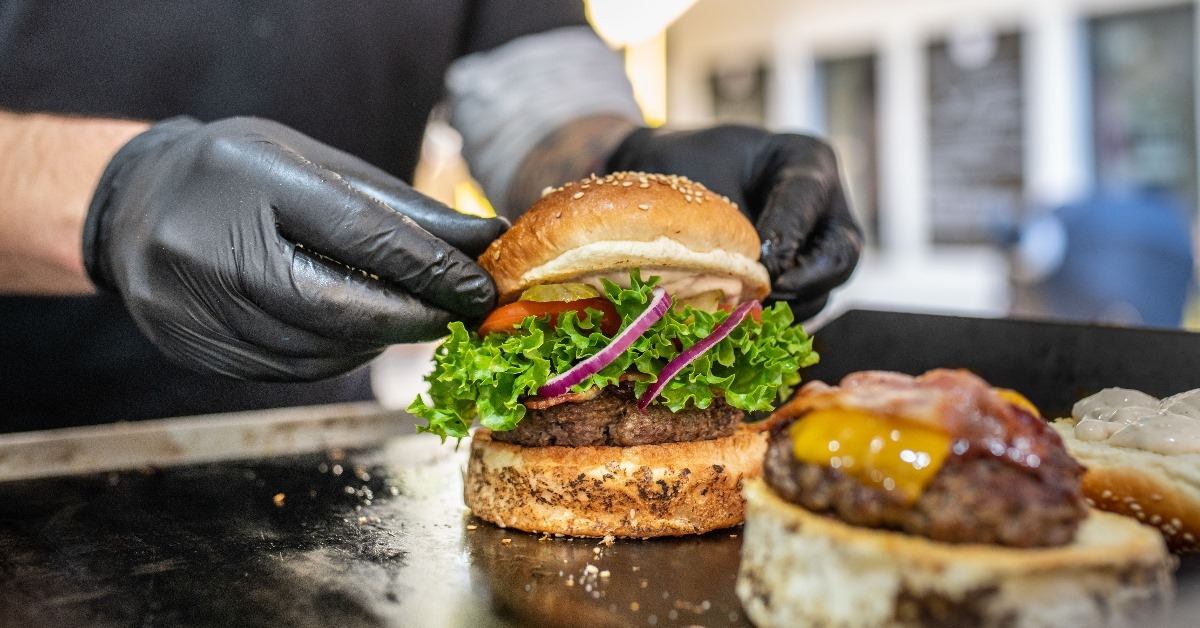 chef wearing black disposable gloves preparing fresh burgers on grill.