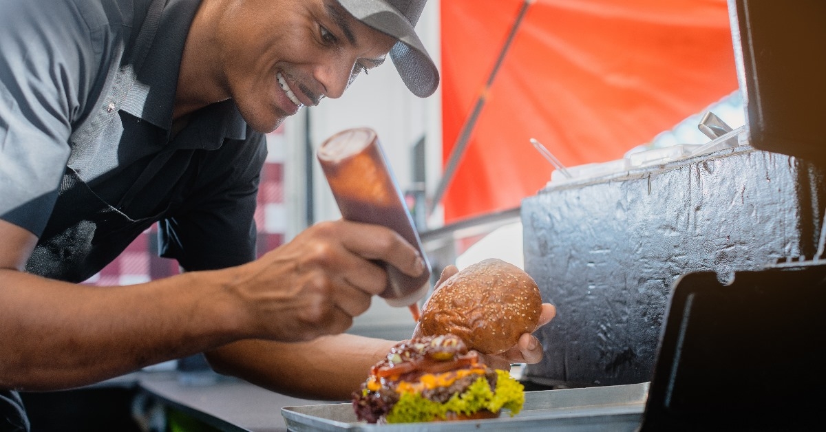 cook in a burger joint finishing a cheeseburger by pouring some sauce over it.