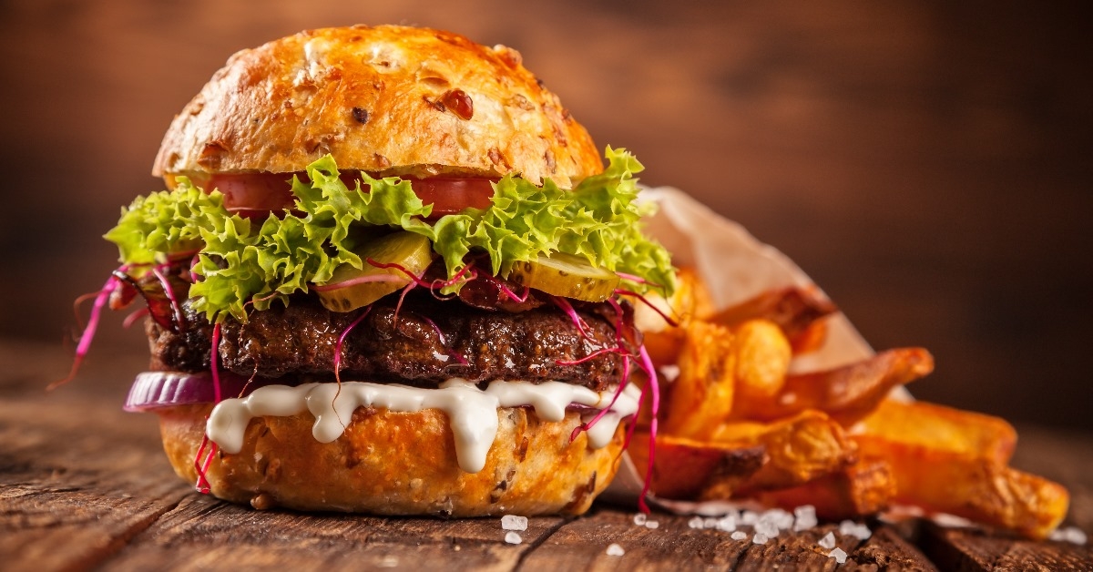 big hamburger with beef patty and sliced pieces of tomato,onion,cucumber served on a big wooden table along with fries. 