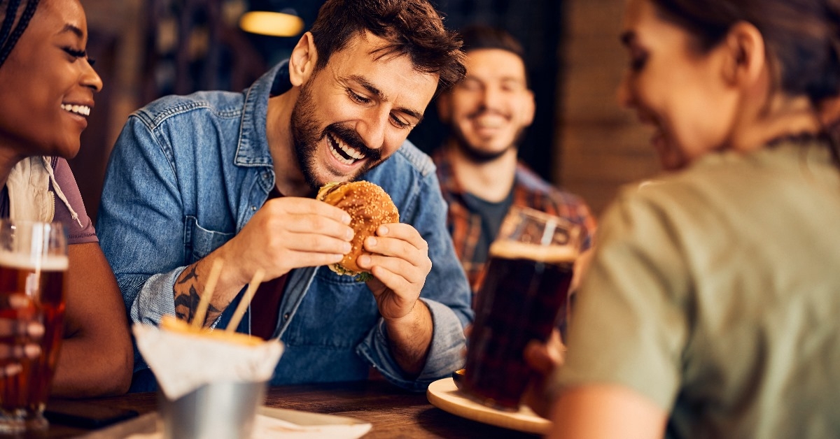 happy man eating burger while gathering with his friends lauging and enjoying drinks in resturant