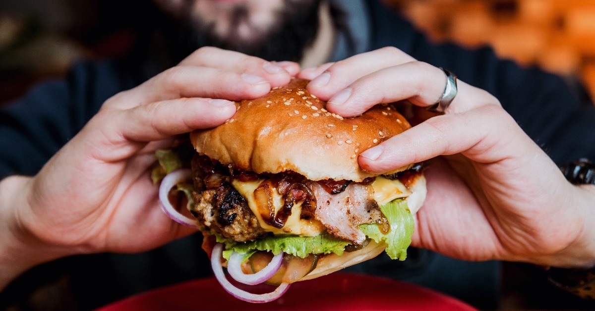 young man eating a cheeseburger with lettuce, bacon and sliced onions