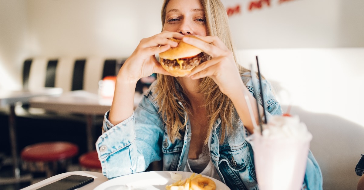 young blonde woman wearing jeans jacket eating burger in restaurant