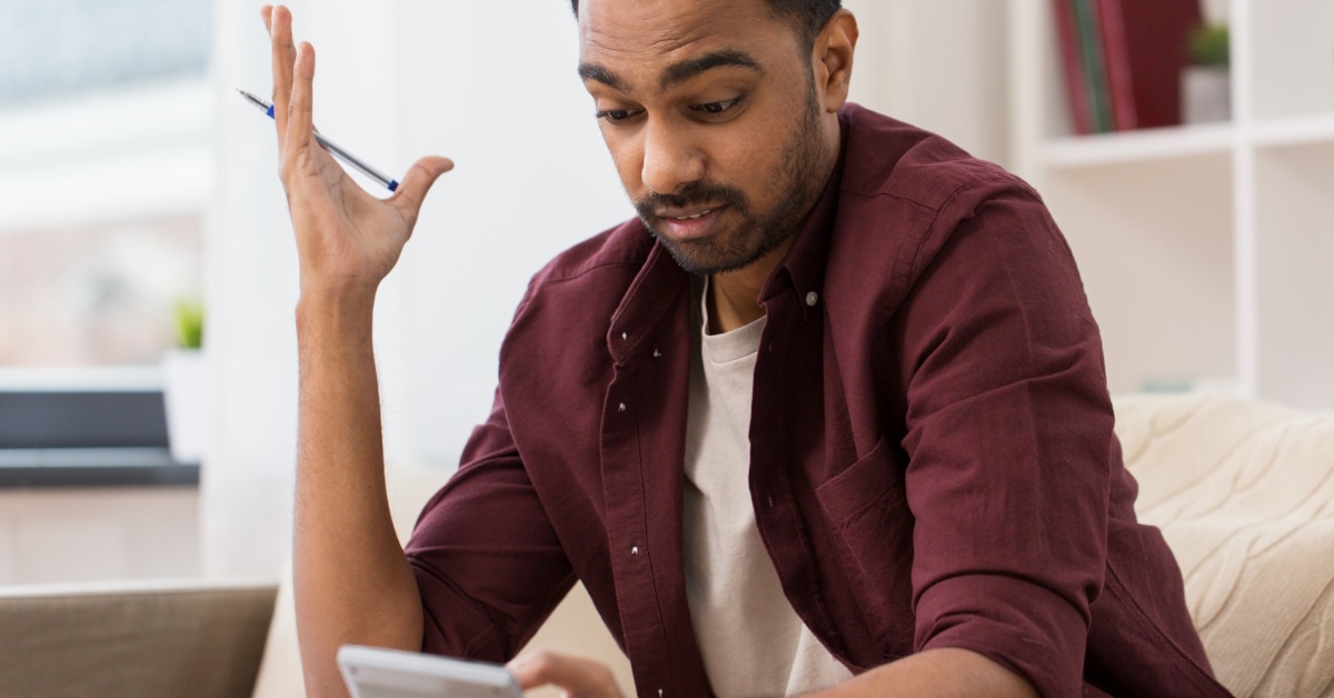 young man sitting on couch at home confused while using calculator