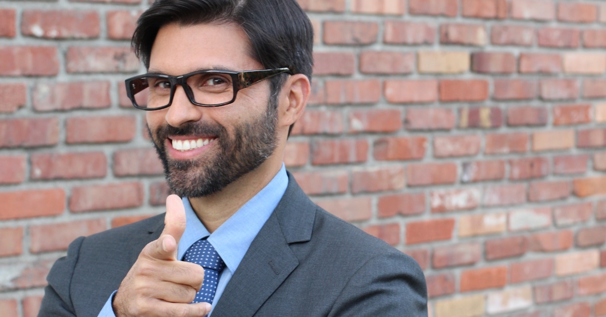 salesman in suit standing over red brick wall smiling while pointing hand towards camera