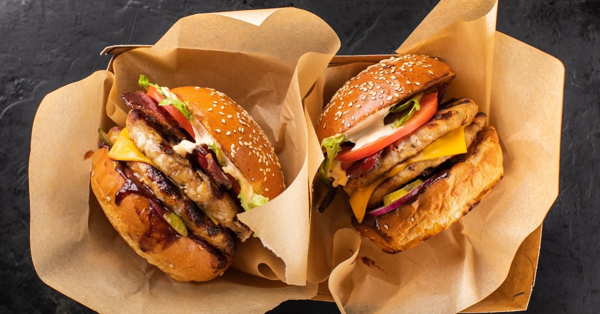 top view of two large takeaway burgers with double chicken patties, cheese, a large slice of tomato and onion. 
