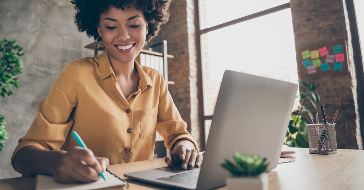 african american woman sitting at table making notes while using laptop at table