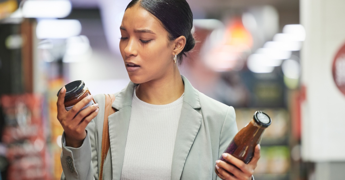 african american woman standing in supermarket reading ingredient label on bottles of sauce