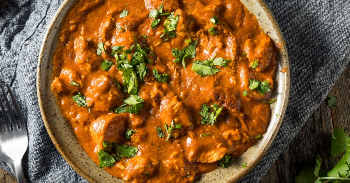 chicken tikka masala in brown bowl with green garnishings on table cloth at wooden table