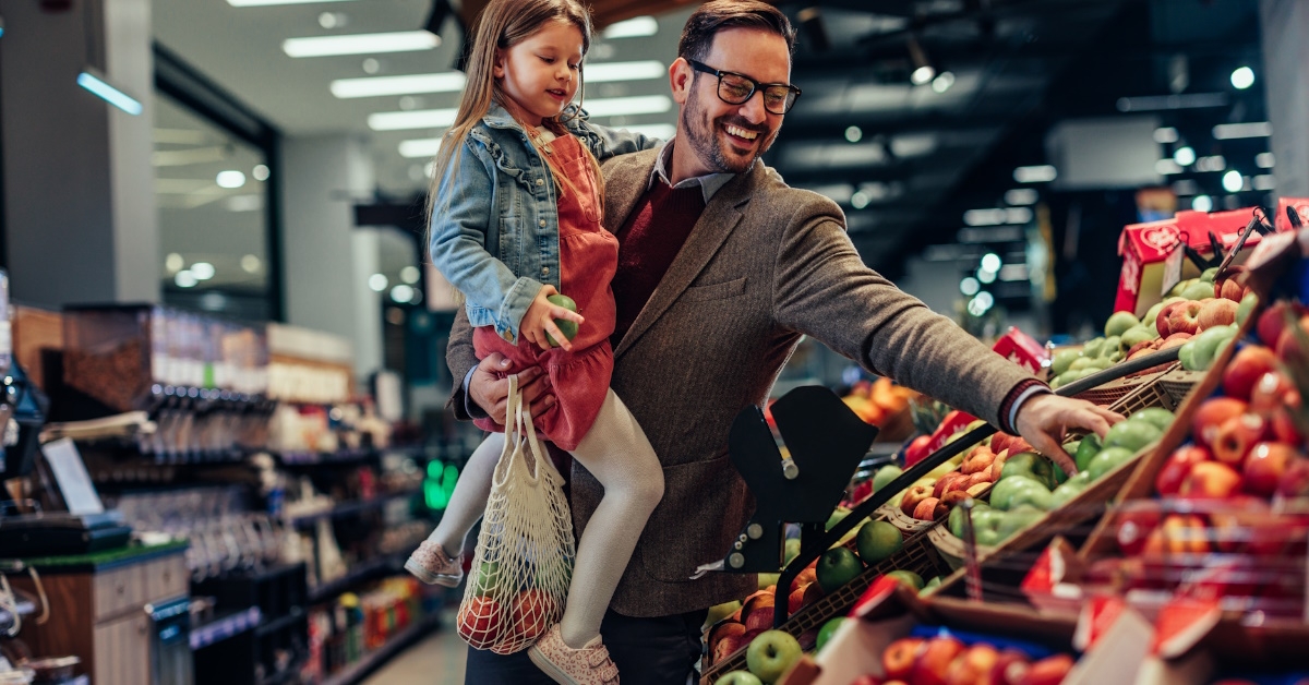 man holding daughter in arms while shopping for red apples in grocery store