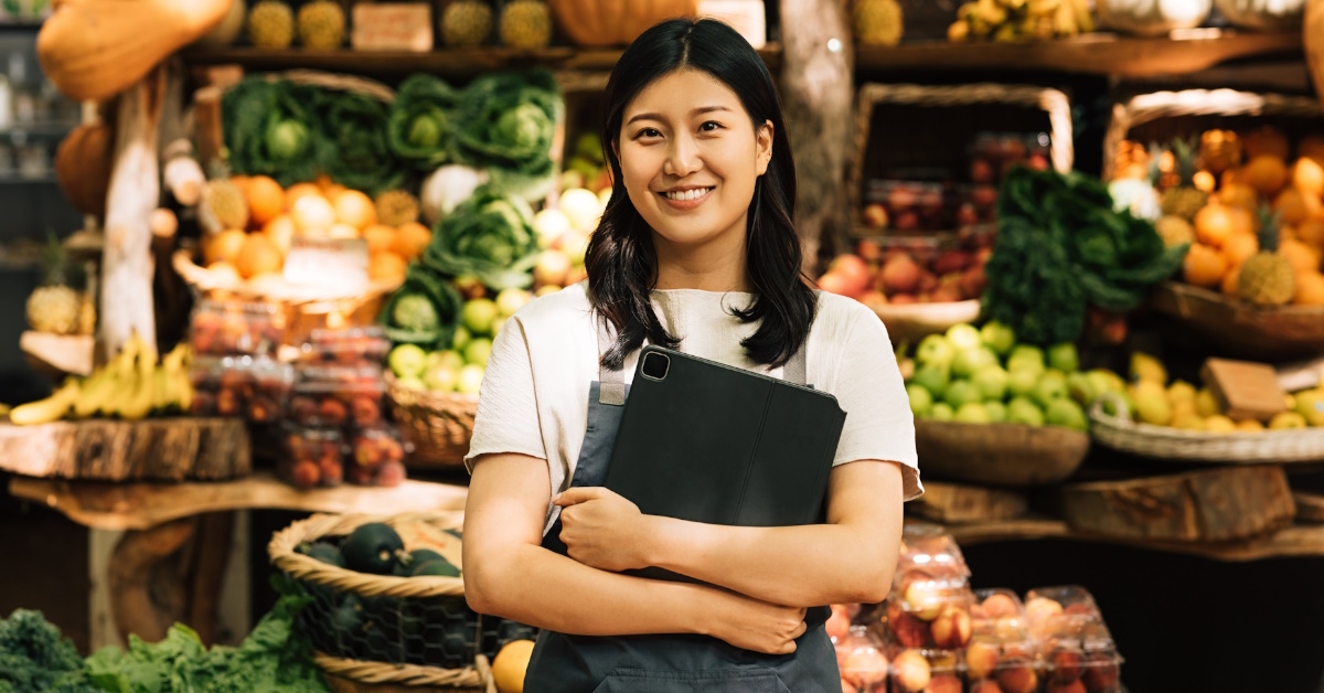 female entrepreneur wearing apron inside grocery store posing for camera while holding digital tablet
