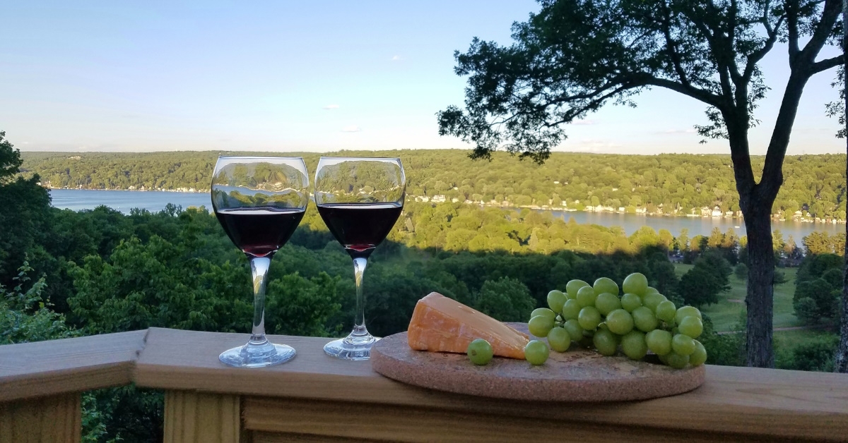  Red wine in glasses with cheese and grapes Against a beautiful view of lake from balcony nearing dusk.