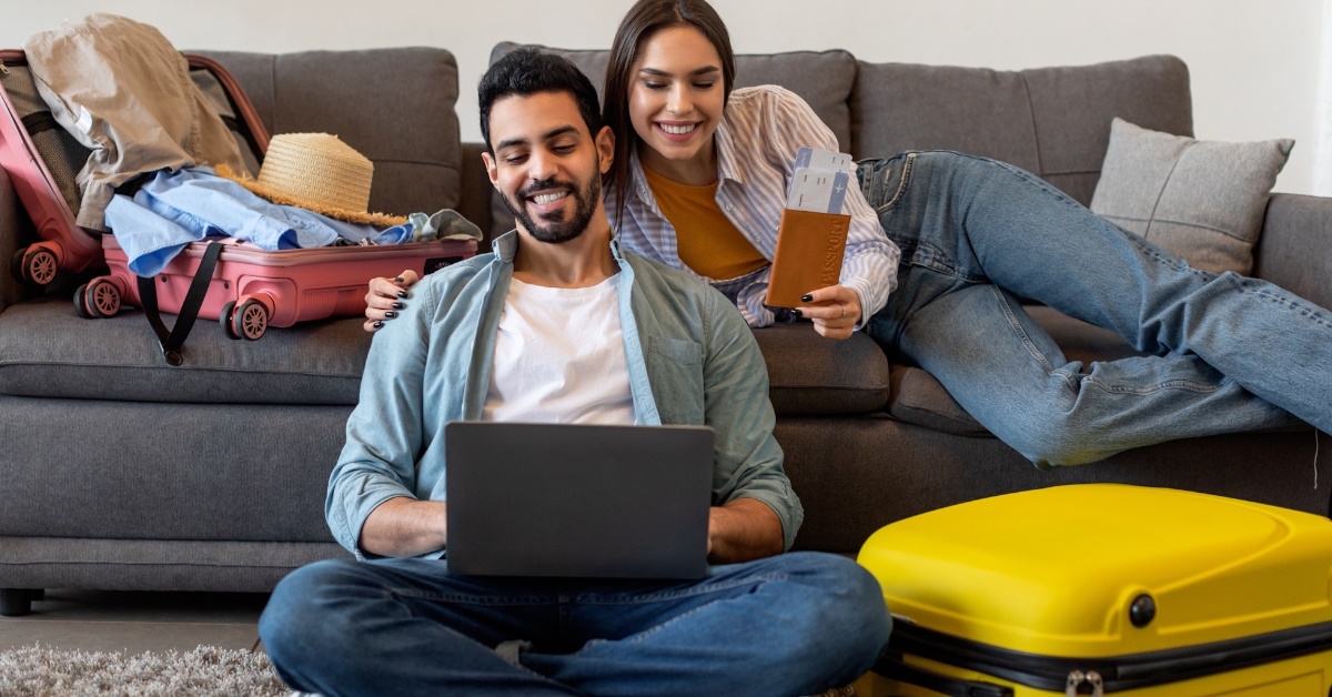Excited couple using laptop to book tickets and hotel online, preparing for vacations and packing suitcases.