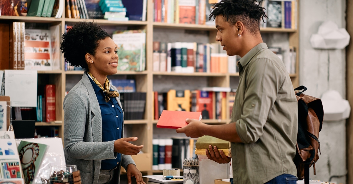 african american boy holding book talking to female librarian in library