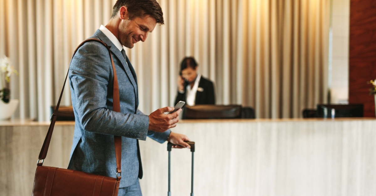 businessman wearing suit and messenger bag smiling using smartphone while walking in hotel lobby with luggage
