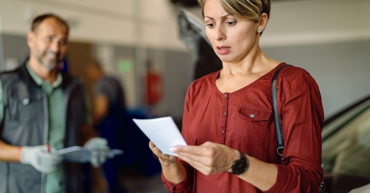 Adult woman holding bill feeling shocked with expense of car repairing in a workshop. Automachanic is standing with his tools in the background.