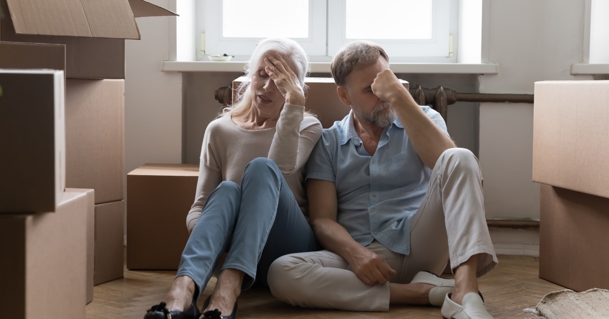 Elder couple tied with relocation, packing and unpacking cardboard boxes, sitting on floor of new home