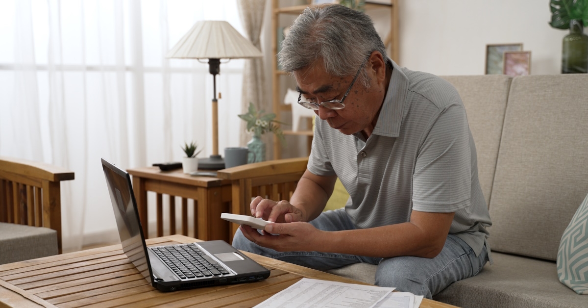 Senior asian man calculating on calculator while sitting in living room sofa using laptop computer and paper indoor
