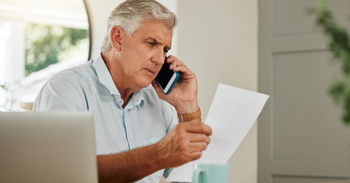 Senior man on a phone call with finance documents and laptop and paperwork checking finances,doing payments.