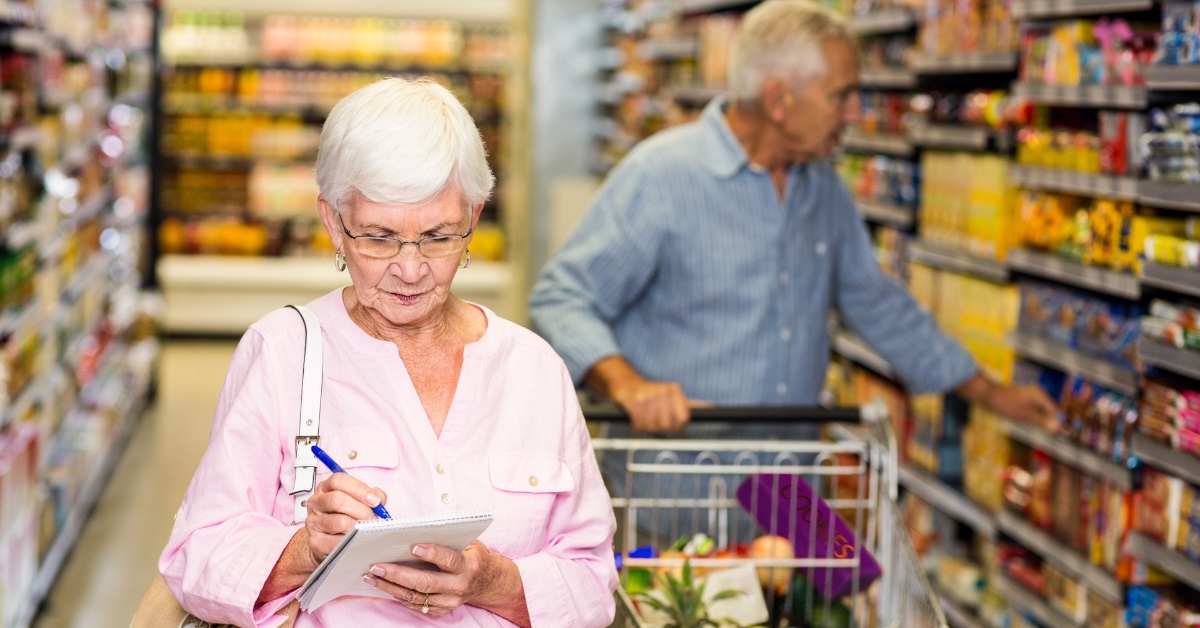 senior women holding a shopping list while his husband is carrying a shopping cart  in background during their shopping in a super store.