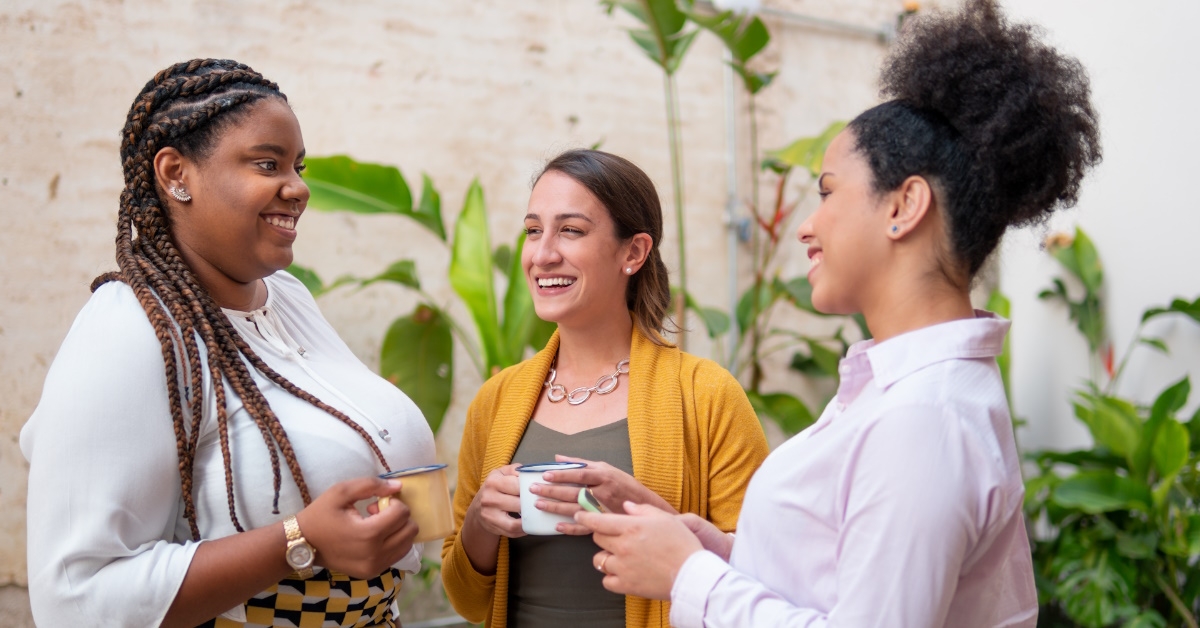 multi ethnic female employees standing outside workplace talking to each other while holding cups and smartphone