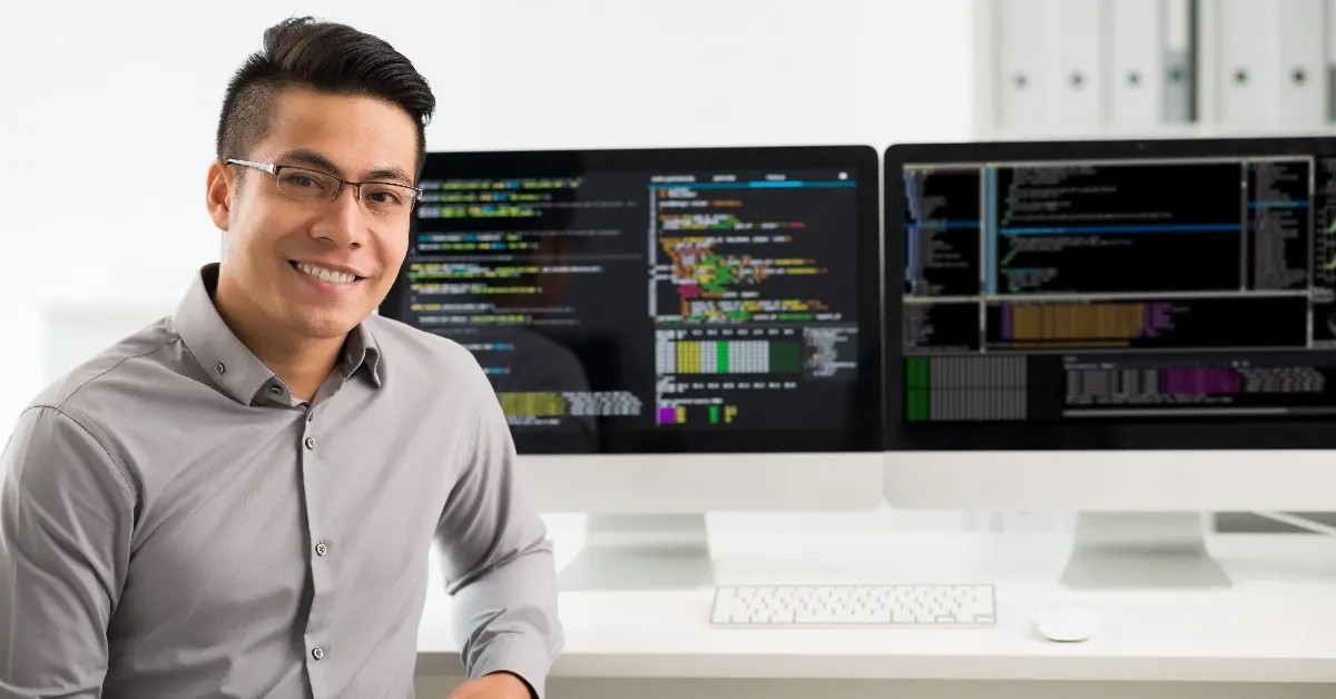 smiling young Vietnamese software engineer wearing formal shirt running programming codes while sitting in front of computer systems.